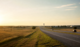 North Texas divided highway at late afternoon through rolling prairie near growing suburbs, with a distant water tower on the horizon