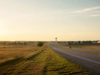 North Texas divided highway at late afternoon through rolling prairie near growing suburbs, with a distant water tower on the horizon