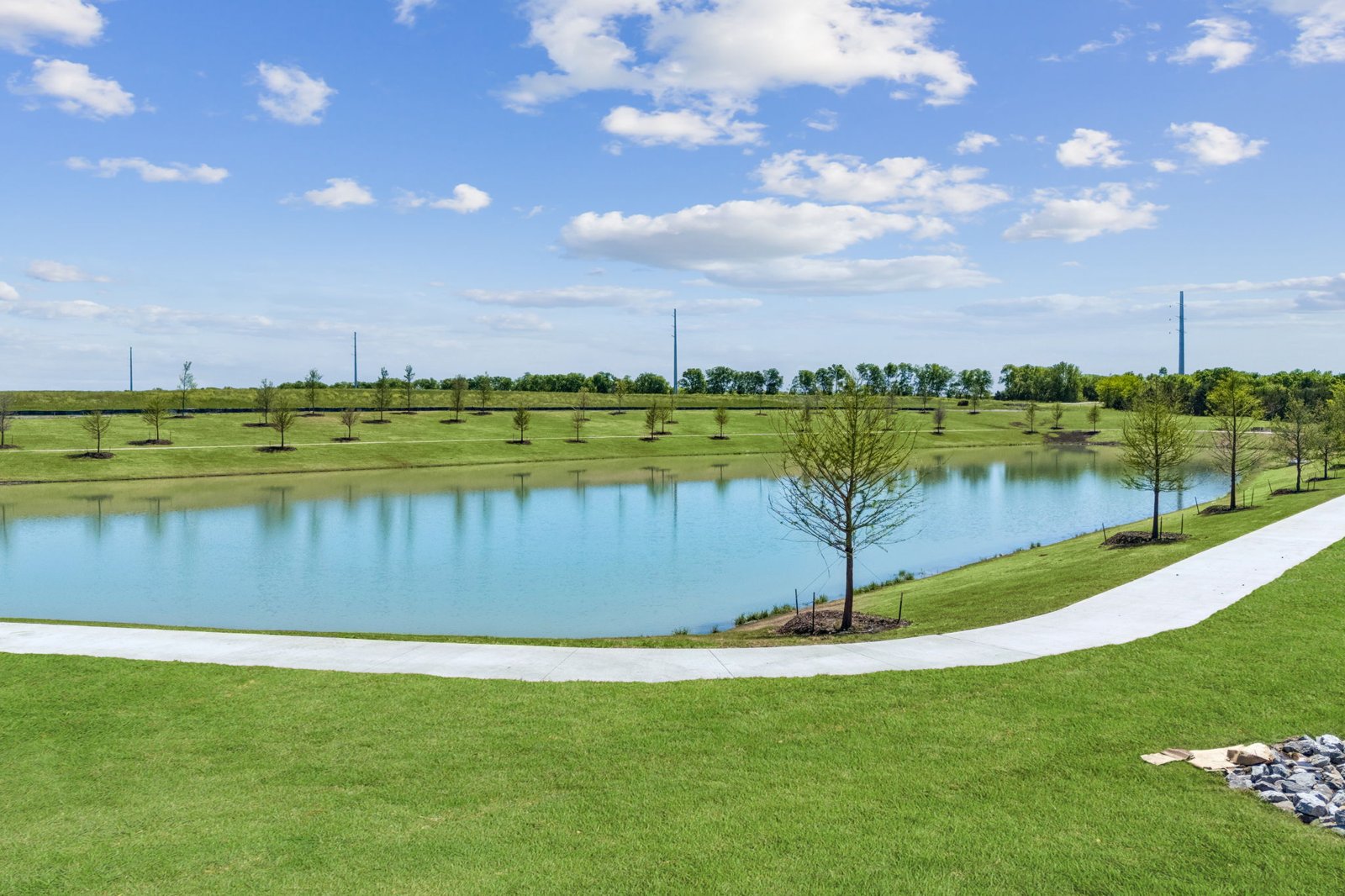Pond with a concrete walking trail and young trees at Sunset Ranch in Gunter TX, with new homes on the far bank